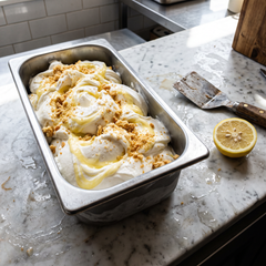 Bacinella di gelato bianco variegato con crema al limone e briciole di biscotto accanto a un limone tagliato