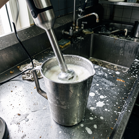 Frullatore a immersione in una caraffa d’acciaio con miscela bianca per gelato a freddo su un banco consumato