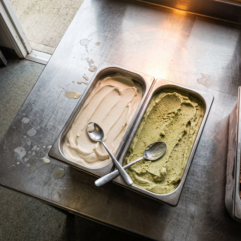 Due bacinelle di gelato vegetale affiancate, una alla soia e una al pistacchio, con cucchiaini d'assaggio