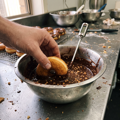 Mano che immerge un donut in una ciotola d'acciaio di glassa al cioccolato croccante con un termometro a sonda, griglia di donut glassati dietro