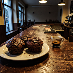 Tre muffin con copertura croccante caffè e cioccolato su piatto in ceramica, sul bancone in legno di un bar vuoto accanto a una tazzina di espresso